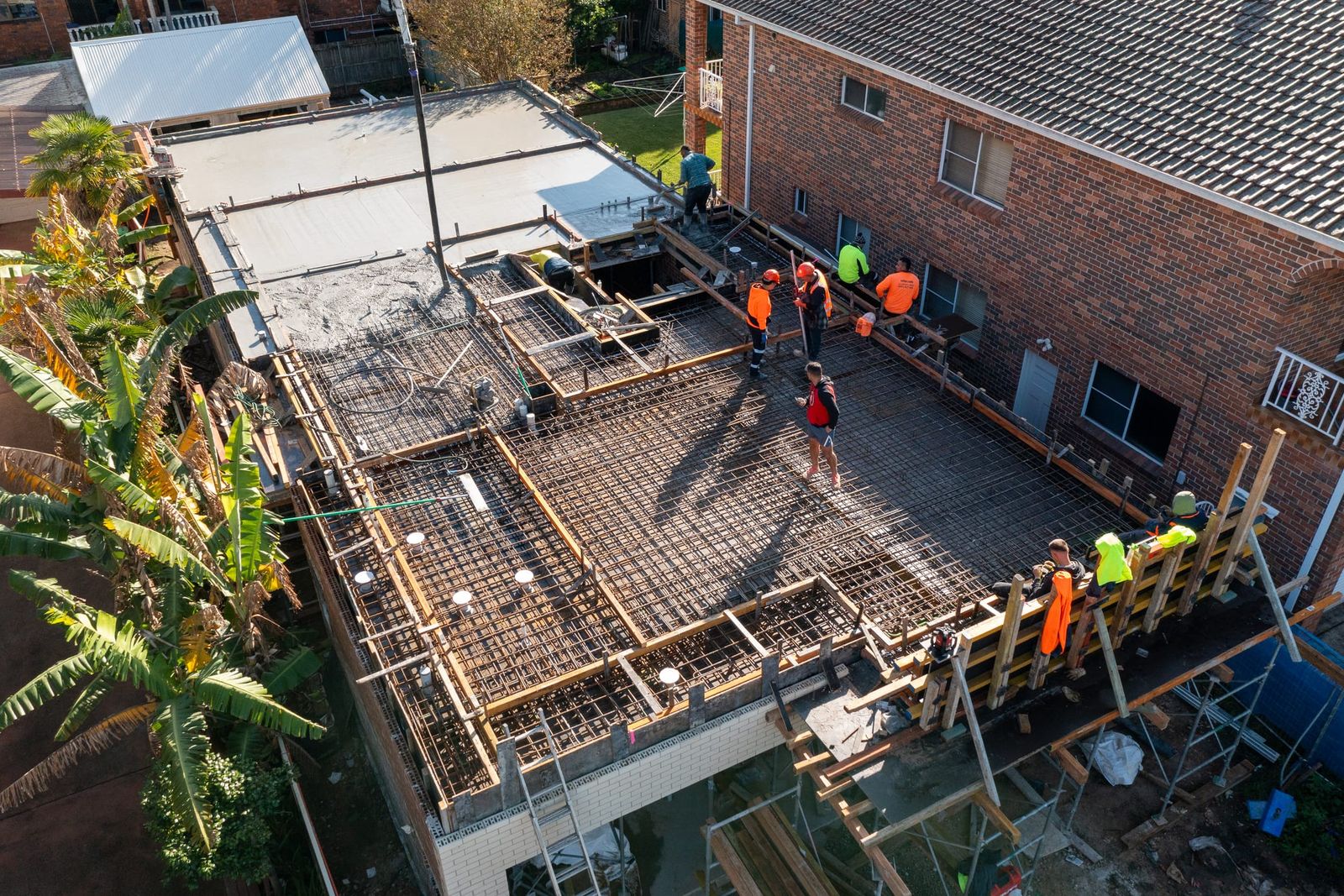 Crew in hi-vis placing concrete over the reinforcement on the suspended slab, a brick neighbour alongside