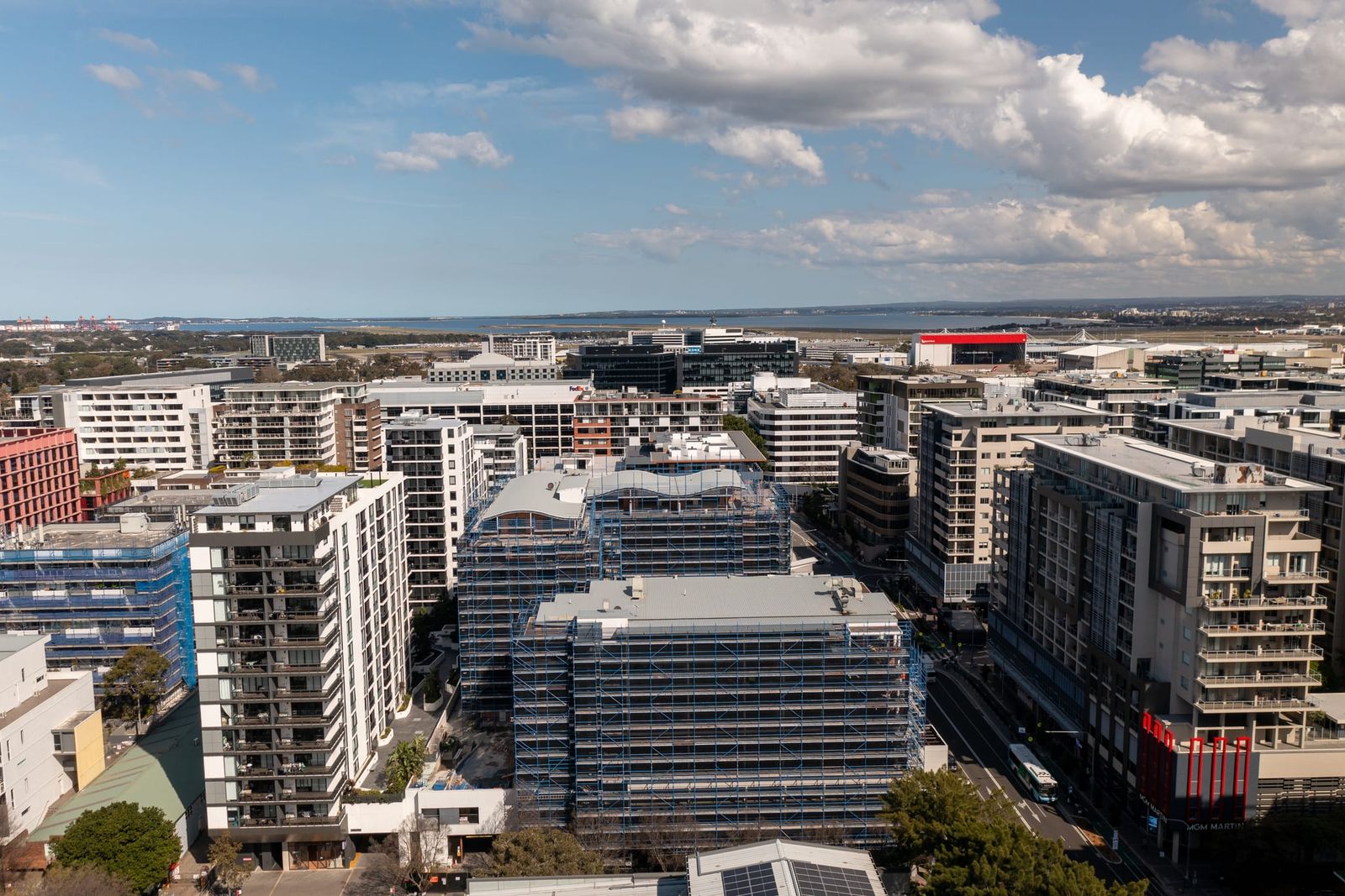 Front-on view of the scaffolded buildings among neighbouring towers, the airport and bay beyond under scattered cloud