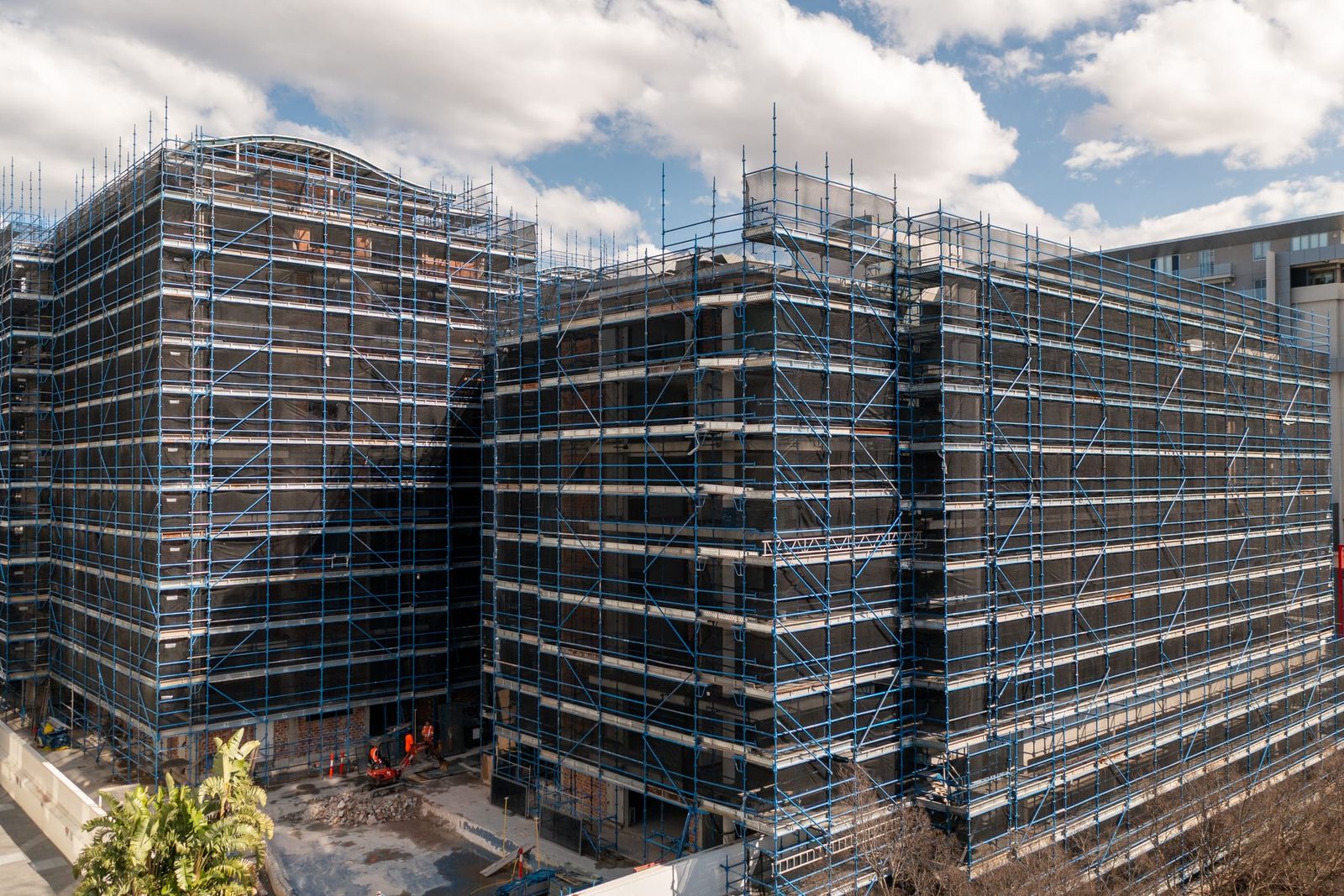 Scaffold across the full width of the buildings, crew in orange at the base under a cloudy sky