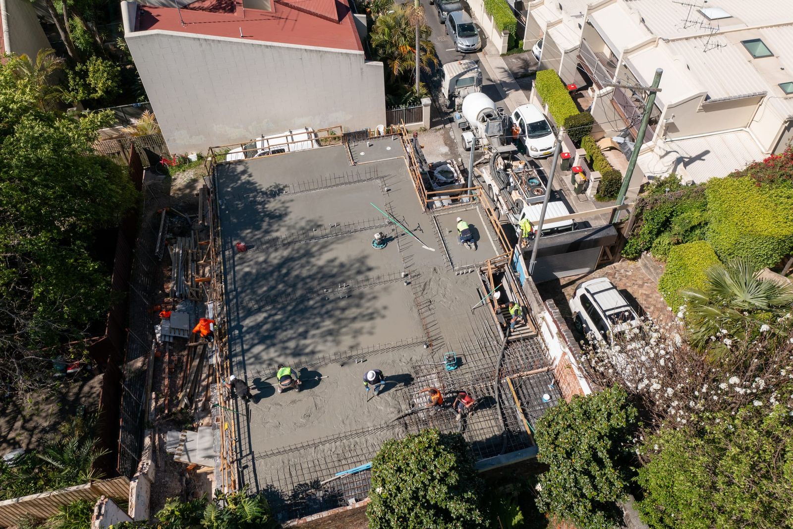 Crew working across the part-poured slab, tree shadows on the concrete and the pump in the lane