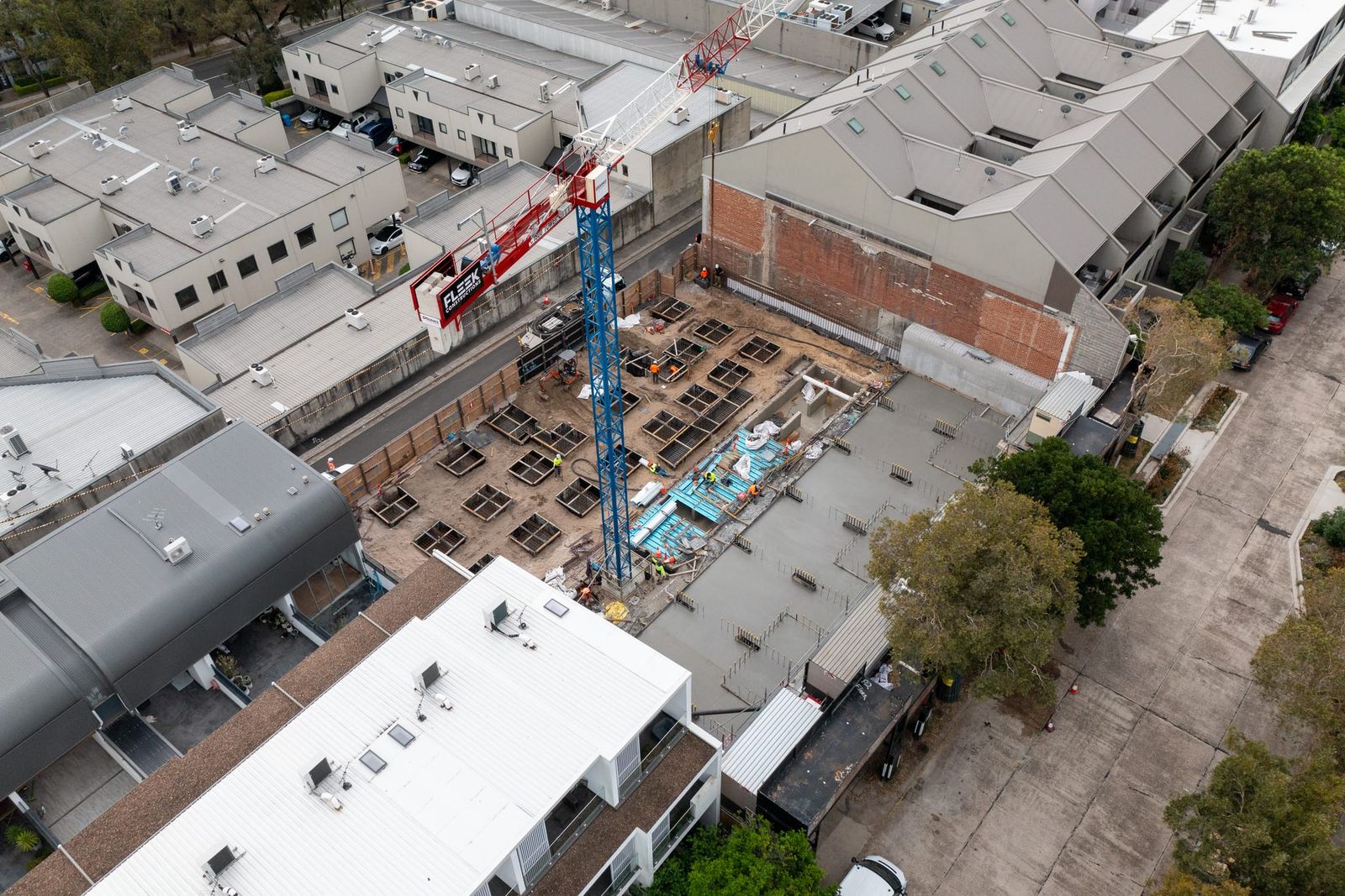 Tower crane standing in the excavated basement, rows of pad footings set out between warehouse roofs and a tree-lined lane