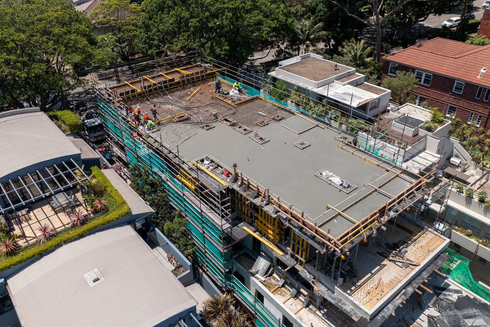 Close aerial of the top deck, formwork crew at the slab edge with scaffold and green mesh down the side of the building