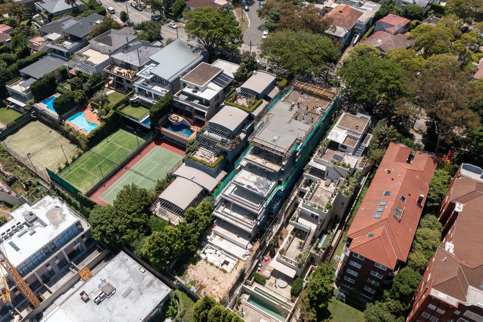 The levels of the build terracing down the slope, green scaffold mesh beside neighbouring tennis courts and flats