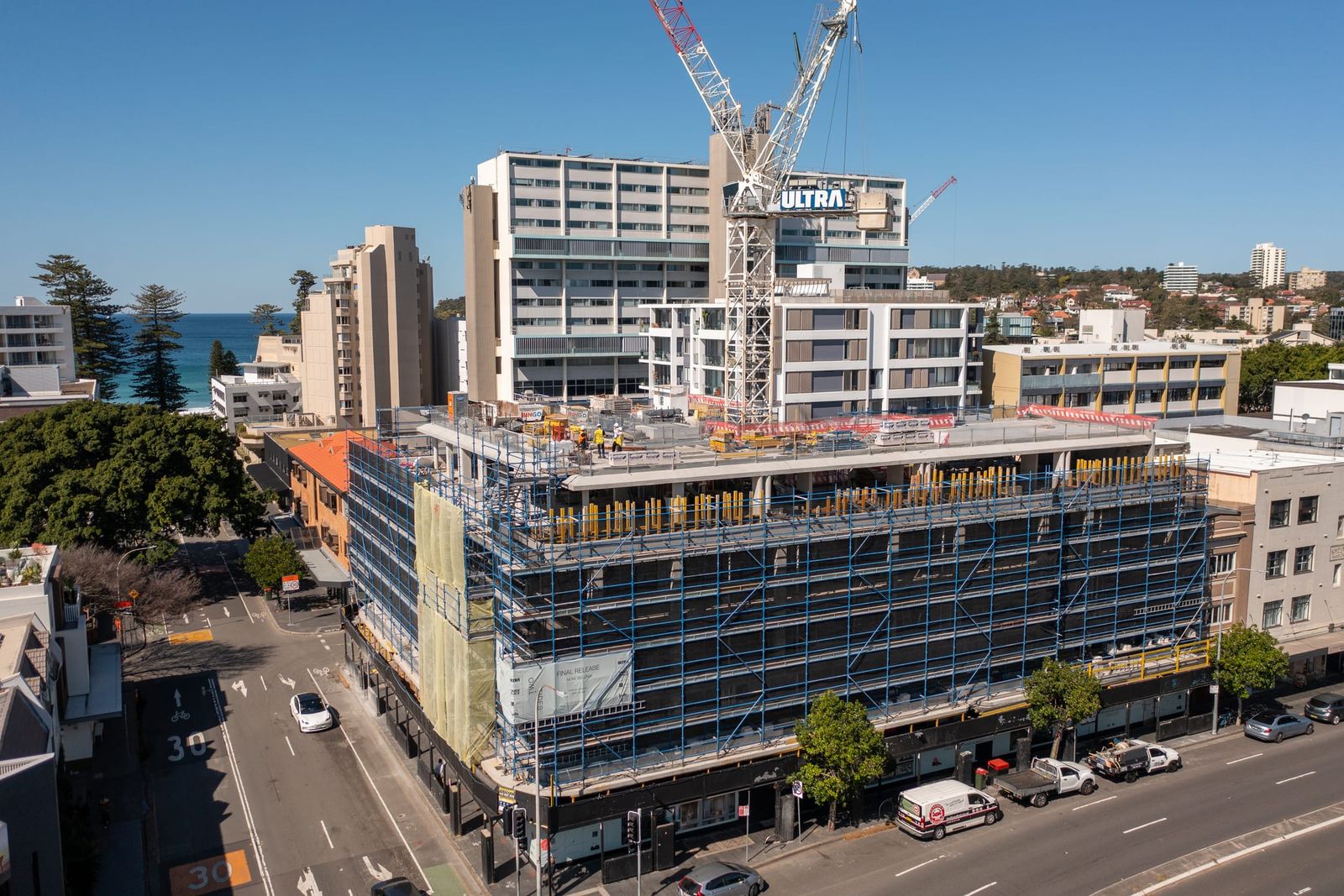Street-corner view of the building wrapped in scaffold, tower cranes above and apartment towers behind