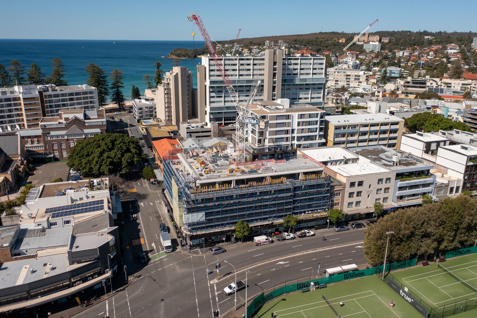Wide view of the scaffolded building beside the tennis courts, the ocean beach and pines beyond