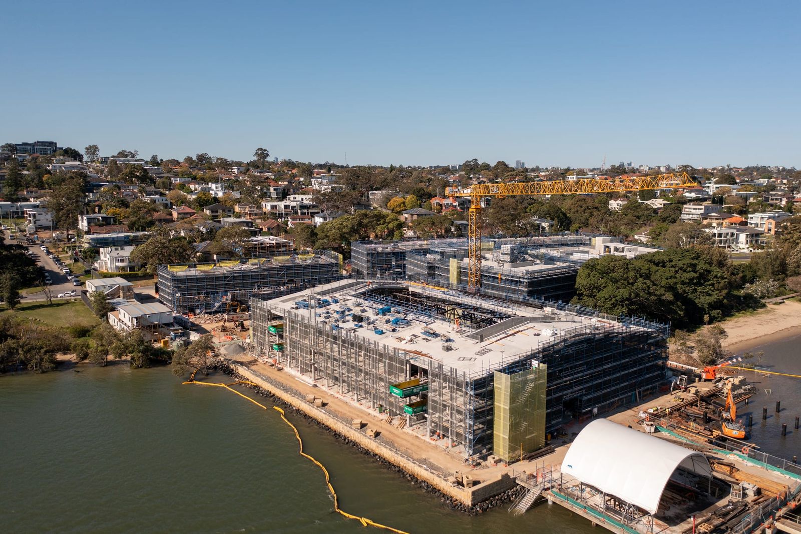Waterfront building wrapped in scaffold, silt barriers along the shore and a white arched structure at the water's edge