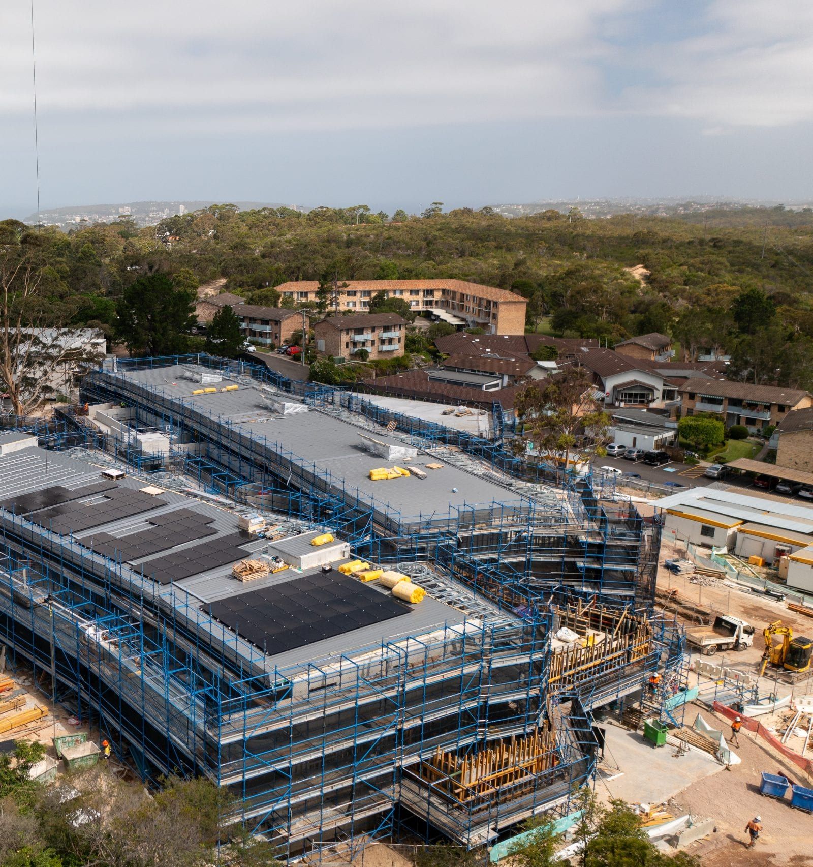 Aerial view of a campus building under construction, wrapped in blue scaffold mesh with roof sheeting underway