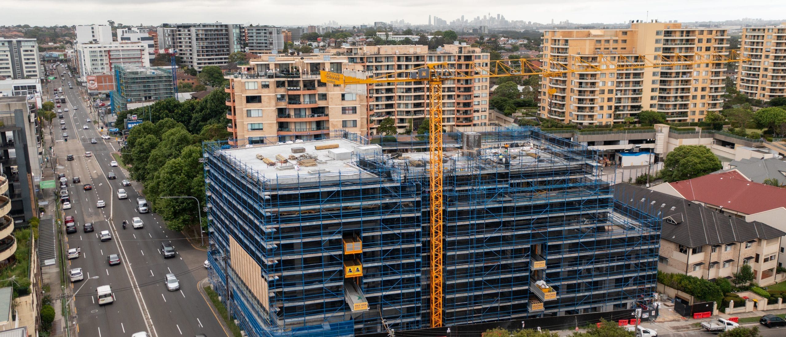 Aerial still of a mixed-use building under construction on a main road, tower crane above, city skyline behind
