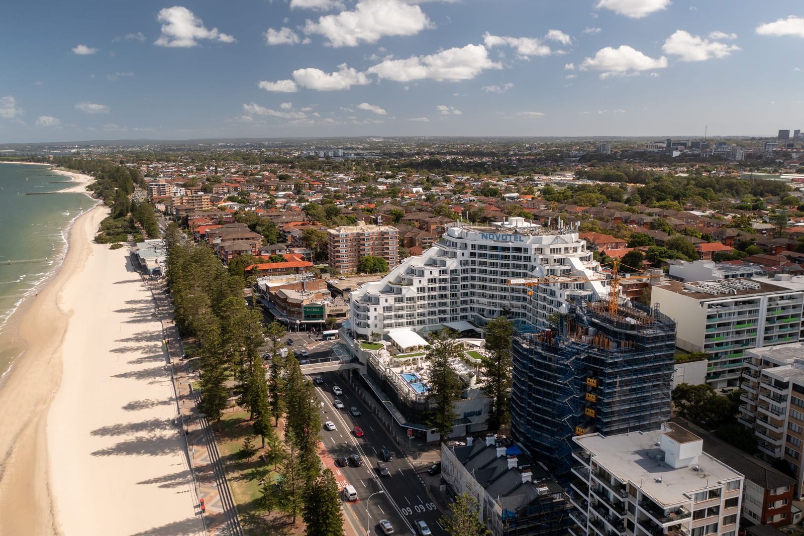 High view along the pine-lined beach, the scaffolded tower beside a large stepped hotel building