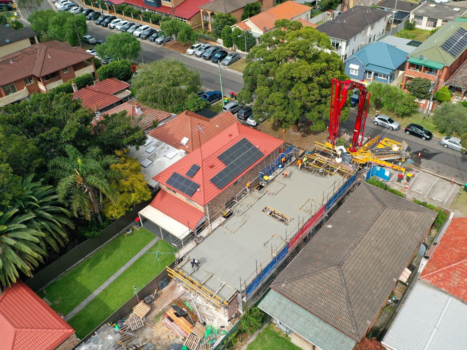 Aerial view of a suburban infill site mid pour, a red concrete pump boom reaching over the fresh slab, crew in hi-vis at the edges and houses on every side