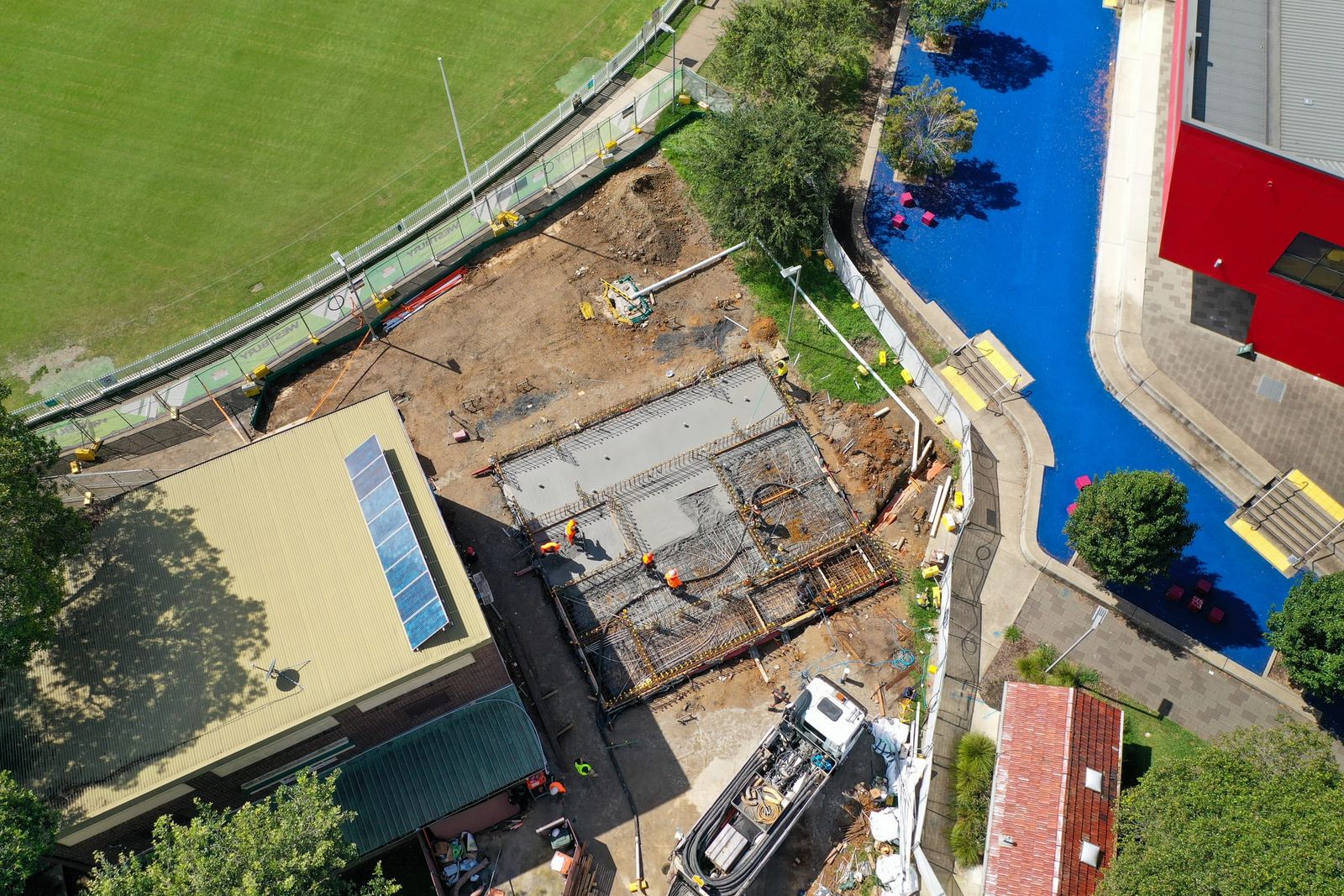 The footings and reinforcement between a brick building and a blue sports surface, a pump truck alongside