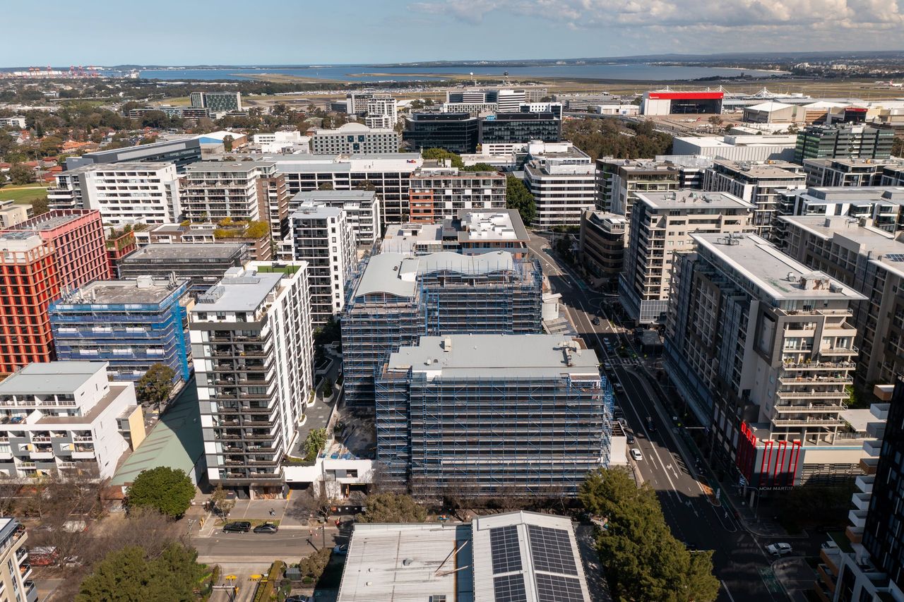 Aerial view over Mascot's apartment precinct, the scaffolded buildings among towers with the airport and Botany Bay on the horizon