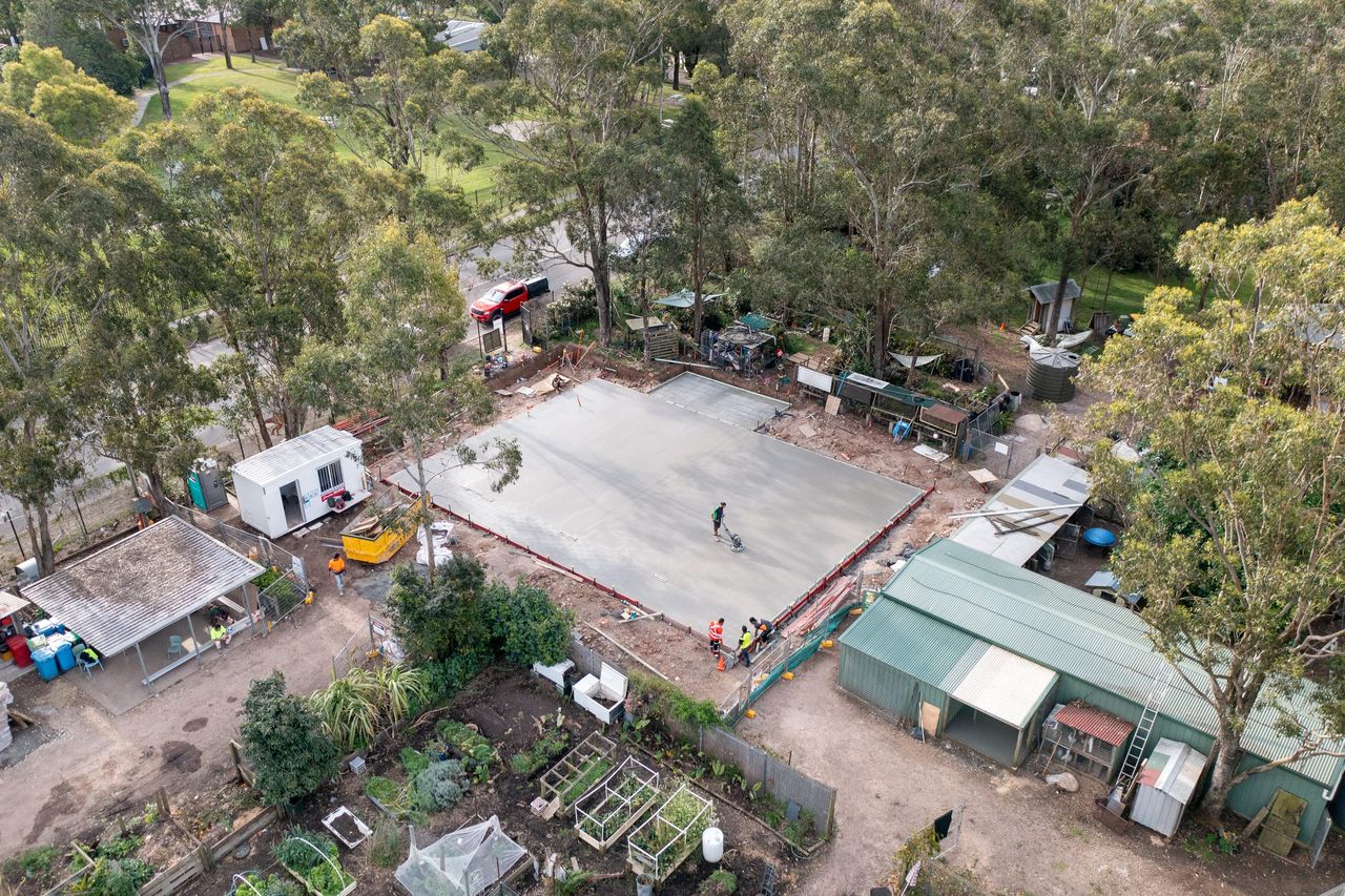 Aerial view of a freshly poured ground slab in Colyton, a worker finishing the surface among gum trees, sheds and a site office
