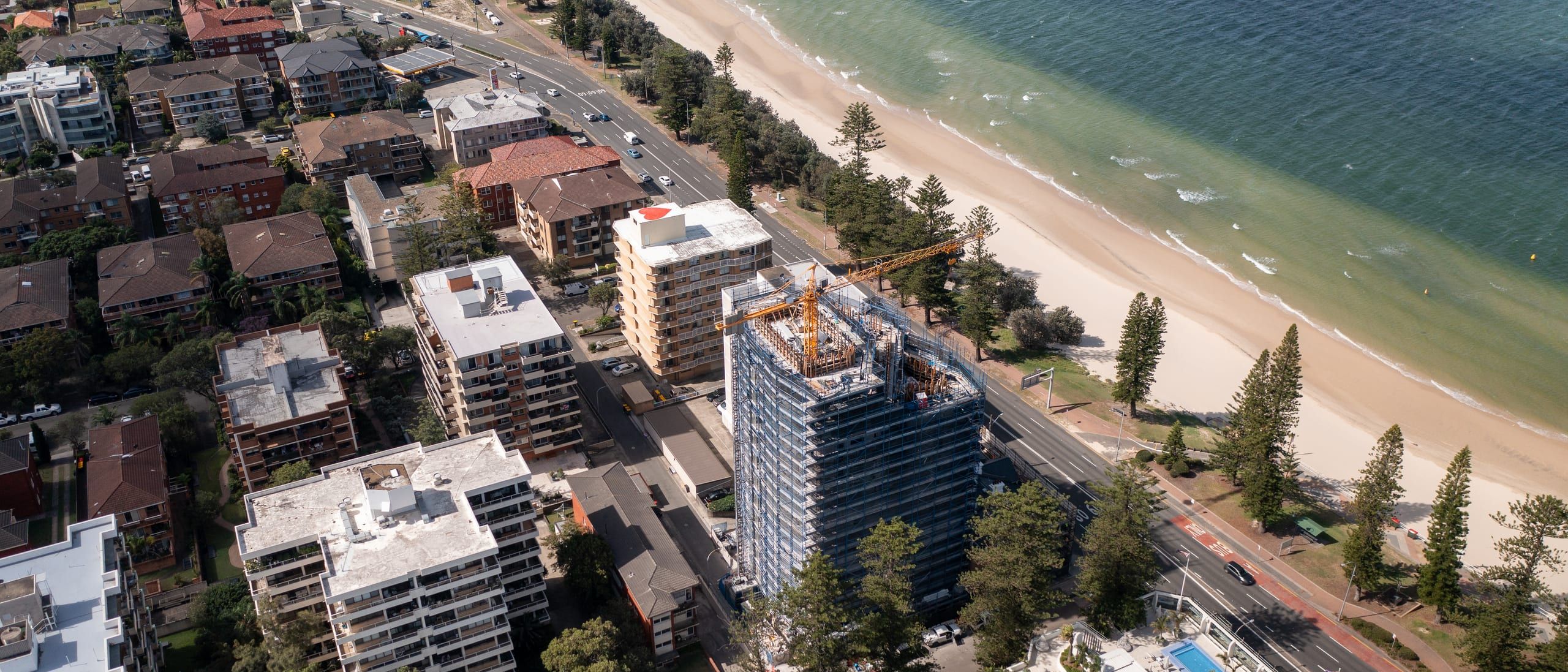 Aerial view along the Brighton Le Sands foreshore, the scaffolded tower beside the beachfront road with Botany Bay alongside
