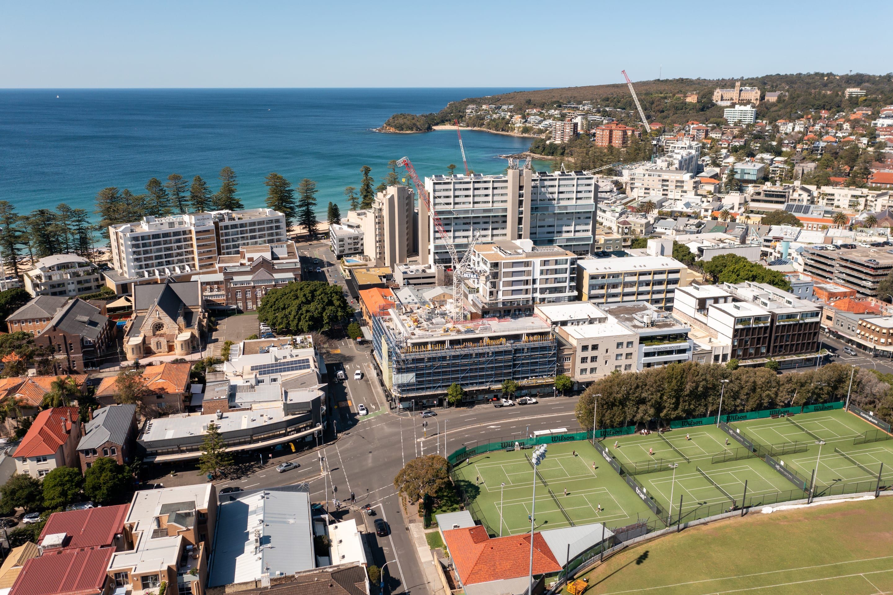 Manly from above, the scaffolded building and crane beside the tennis courts, the ocean beach and headland behind
