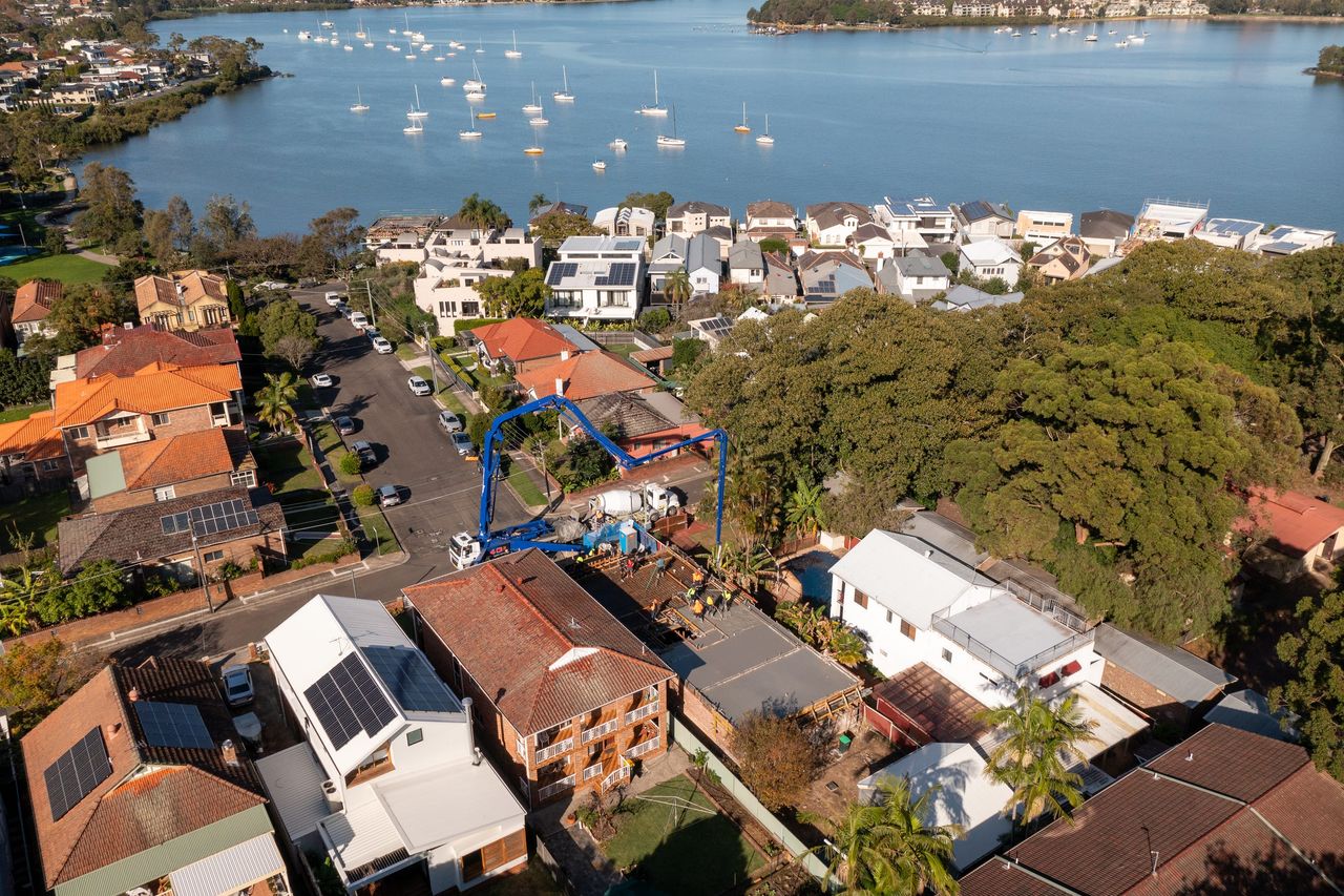 Aerial view over Abbotsford to the Parramatta River, a blue boom pump reaching over a home under construction, moored boats on the bay