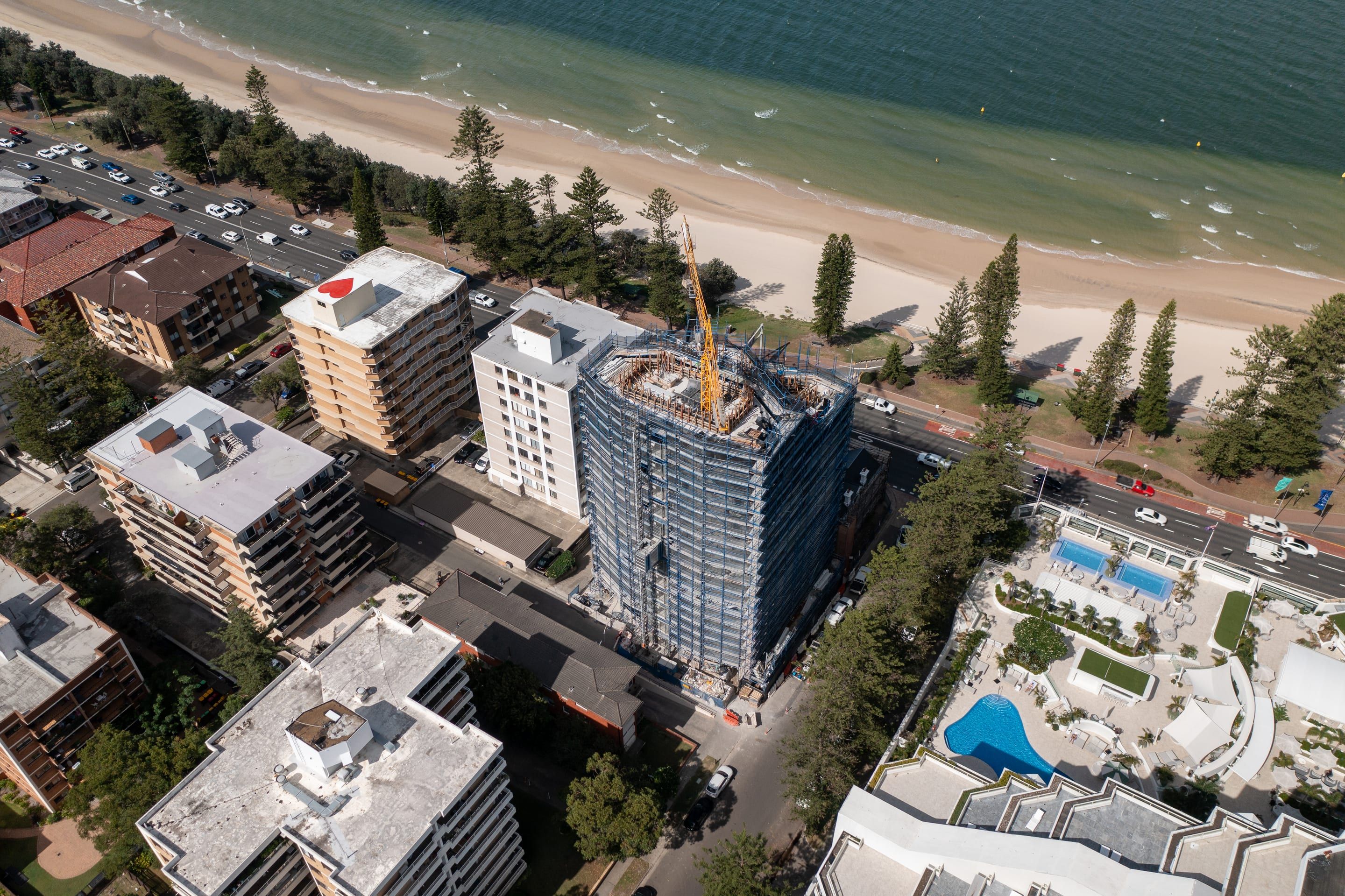 The tower wrapped in scaffold from ground to roof with a tower crane on top, between apartment blocks and the Norfolk pines