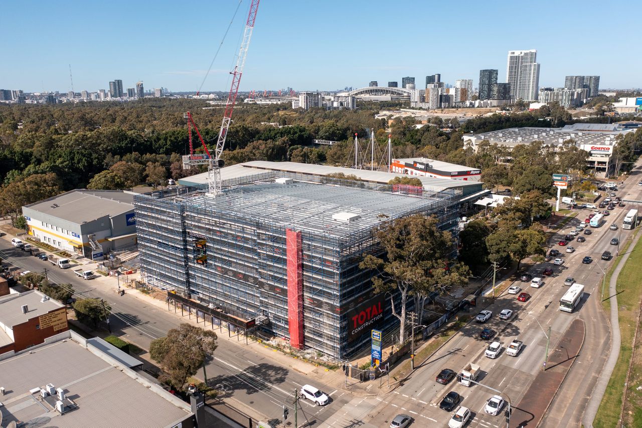 Aerial view of the scaffolded building on a busy road in Lidcombe, a tower crane above and the Sydney Olympic Park towers on the skyline